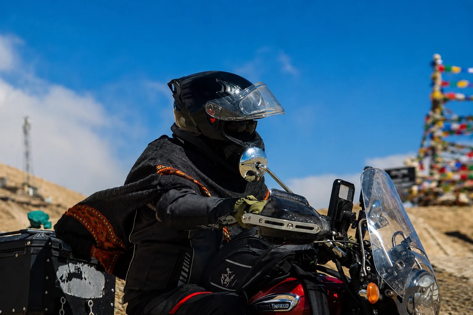 A helmeted motorcyclist in a traditional poncho riding a Royal Enfield in the Himalayas